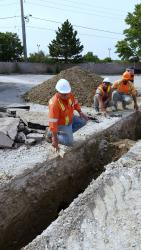 Worker in orange jacket and white helmit looking in trench.
